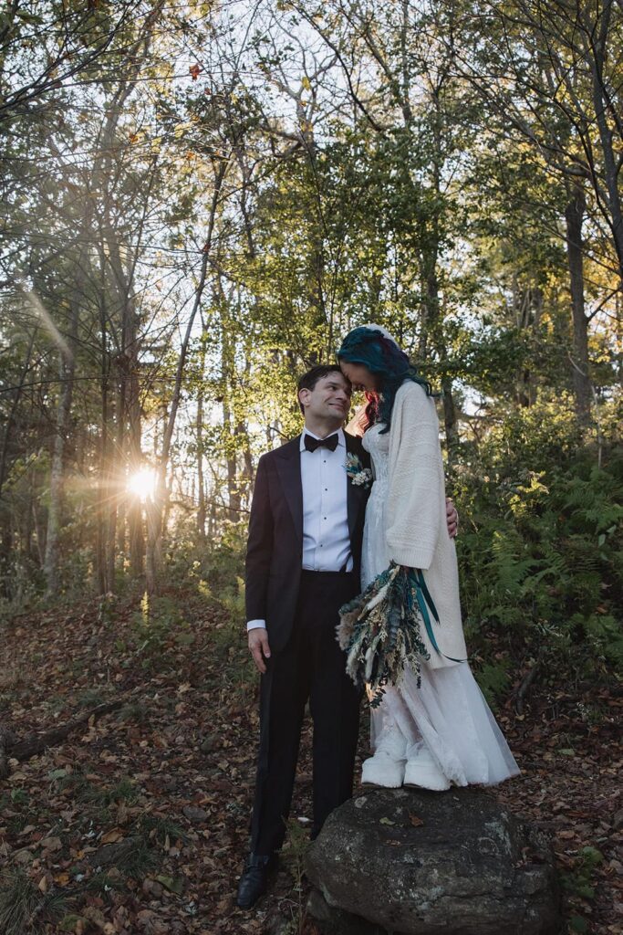 A bride stands on a rcok to be taller than her husband and leans her forehead against his. The sun is rising behind them