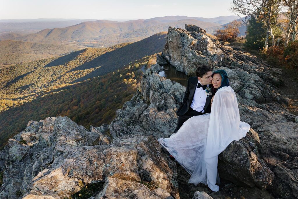 A bride and groom sit on the rocks at Little Stony Man during the adventure elopement at Shenandoah. The groom is kissing the cheek of his bride. He is wearing a tux and she is wearing a dress with a sheer cape.