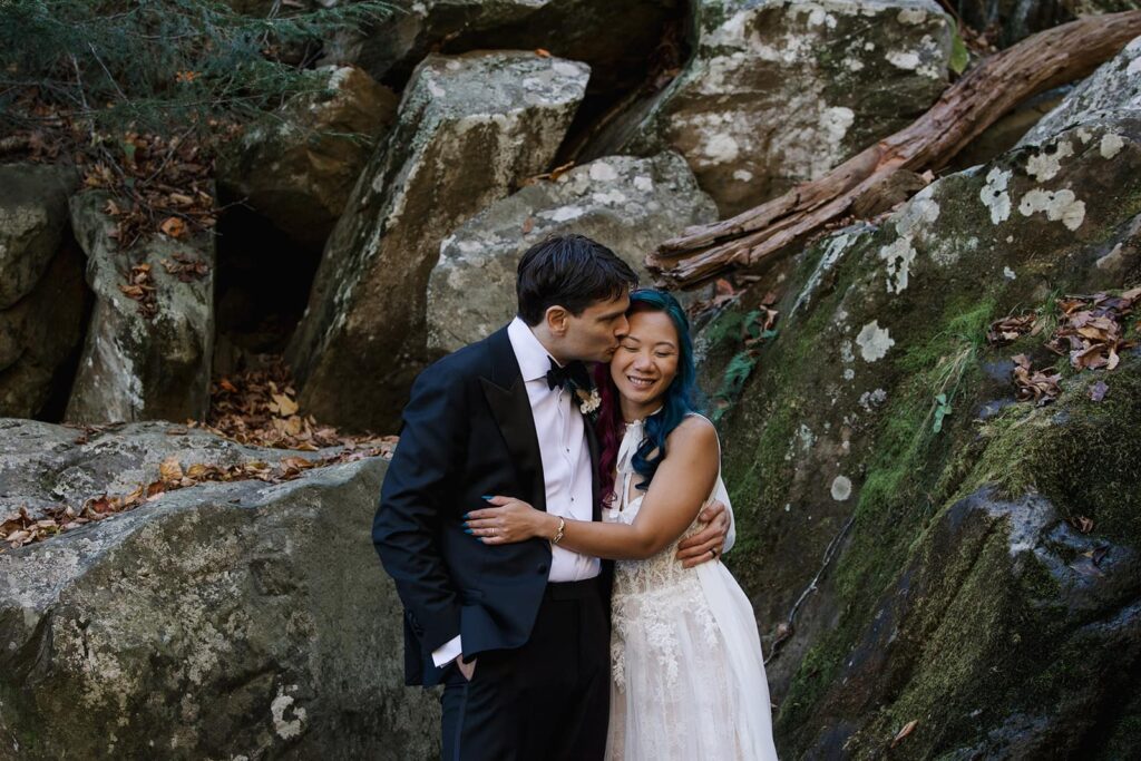 A groom kisses the cheek of his bride, and she is smiling. They are standing in front of some boulders near Dark Hollow Falls