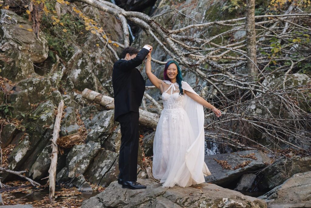 A groom twirls his bride in front of Rose River Falls while she laughs. 