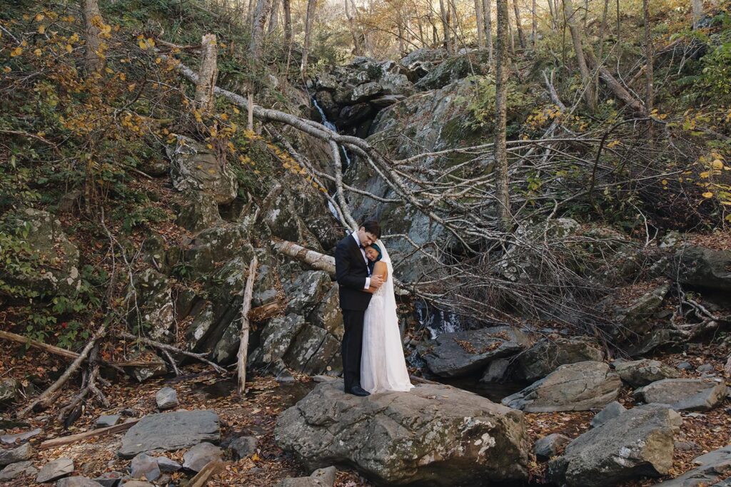 A bride and groom embrace while standing on some giant boulders in front of Rose River Falls  in Shenandoah on their elopement day