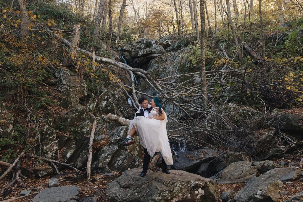 A groom lifts his bride and spins her while standing in front of Rose River Falls in Shenandoah during their adventure elopement.