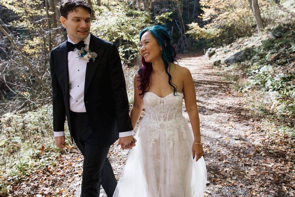 A bride and groom walk while holding hands on a sunlit trail in Shenandoah. The groom is wearing a tux and the bride is wearing a lace strapless dress