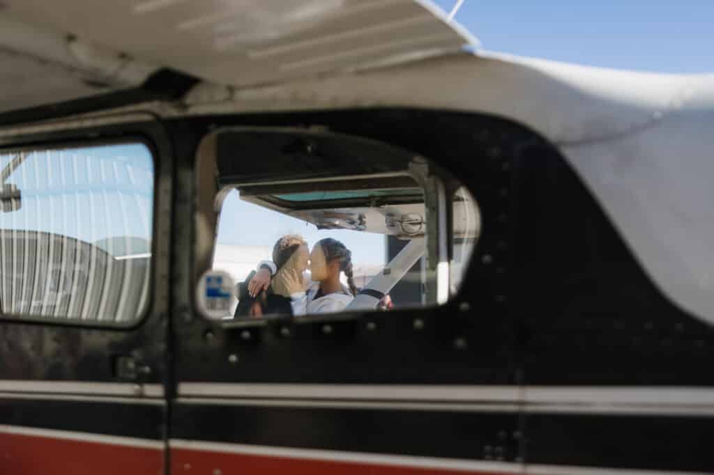 A photo taken through the window of a small plane where you can see a bride and groom kissing on their wedding day