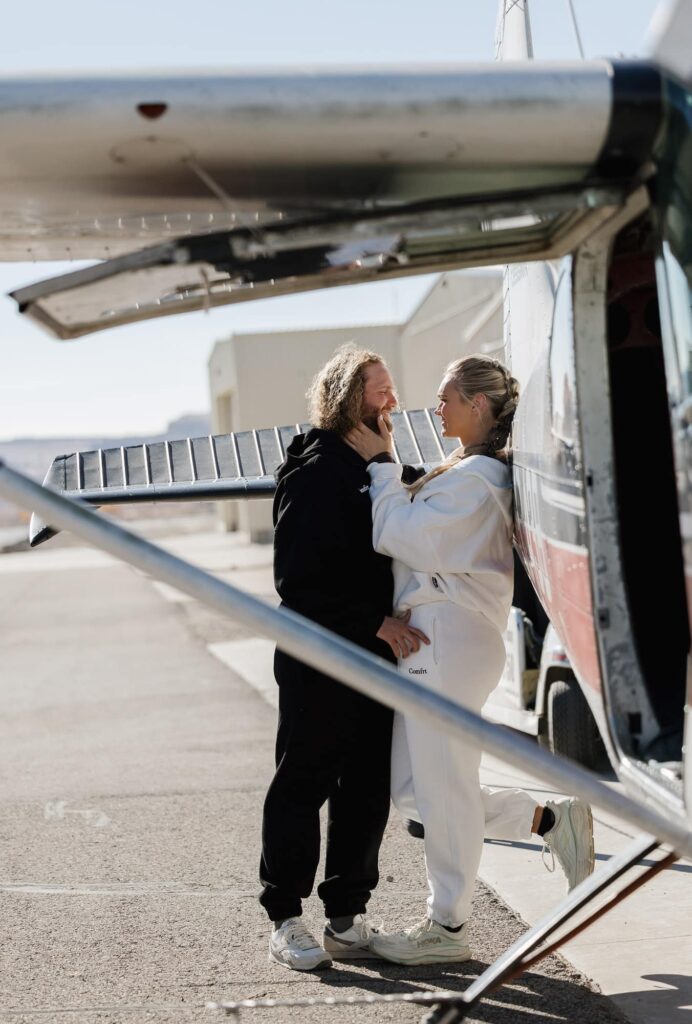 A bride is leaning against a plane, while the groom leans in towards her. The couple is waiting to go skydiving on their elopement