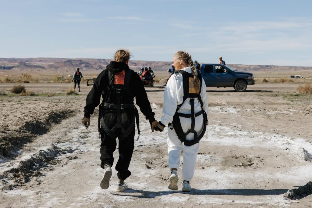 A bride and groom walk holding hands still in their skydiving harnesses after skydiving on their wedding day