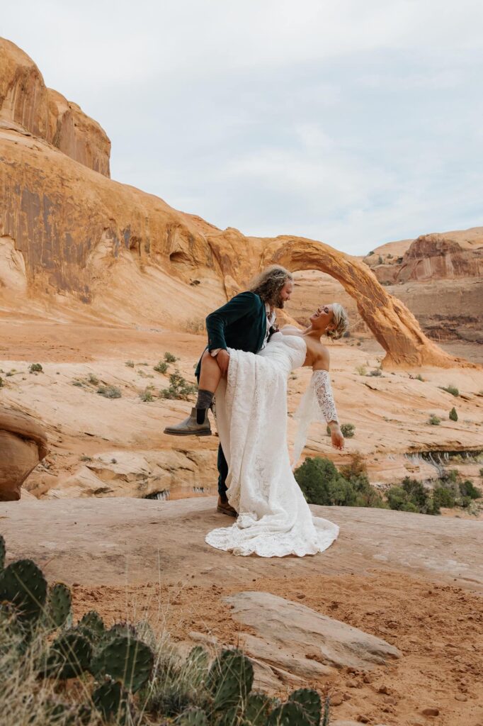 A groom dips his bride in front of the Corona Arch during their adventure elopement in Moab