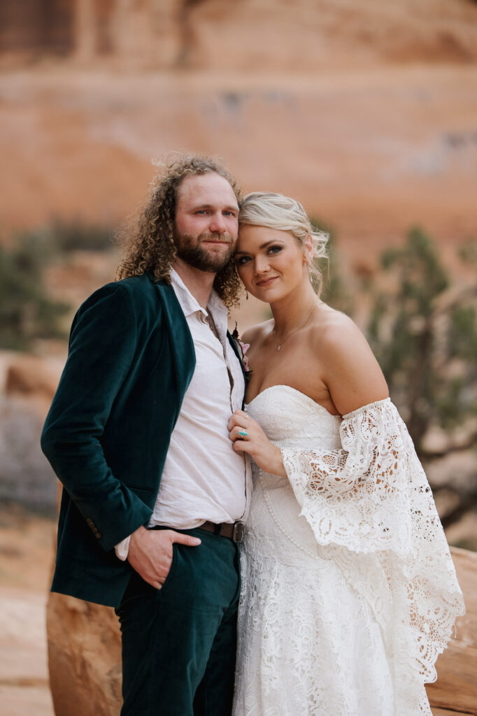 A portrait of a bride and groom on their wedding day. The groom is wearing a green velvet suit and the bride is wearing a romantic boho dress with lace sleeves