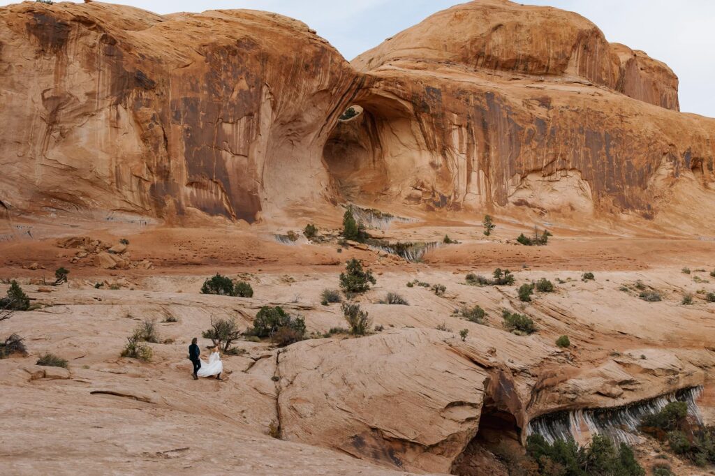 A bride and groom hike towards the Corona Arch in Utah
