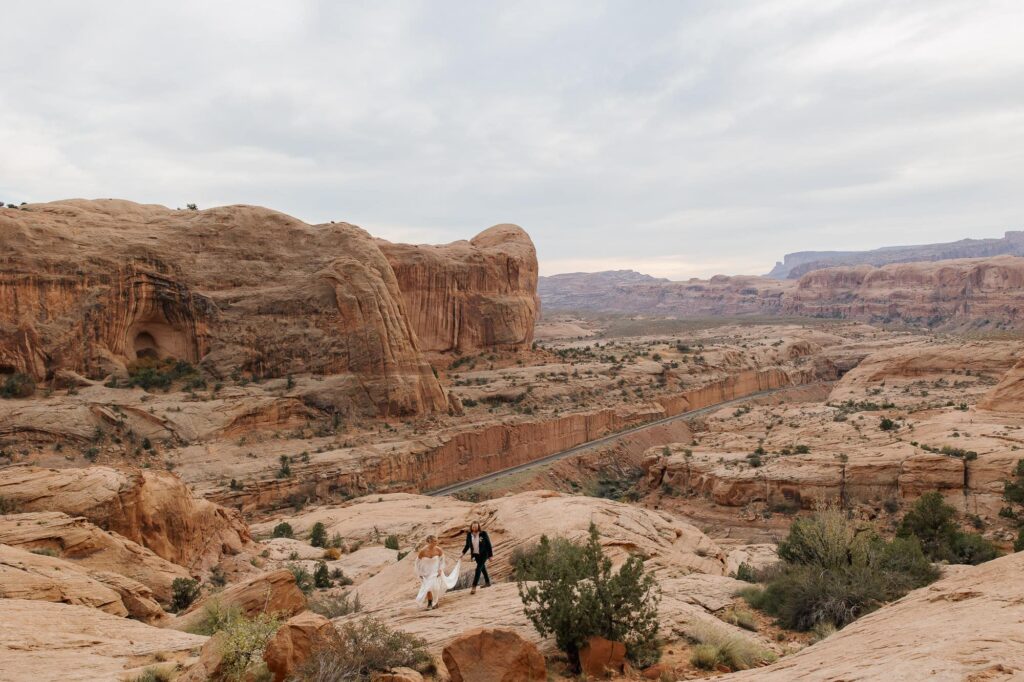 A bride and groom walking in a desert landscape in Moab. The groom is holding the dress of his bride so she can walk. 