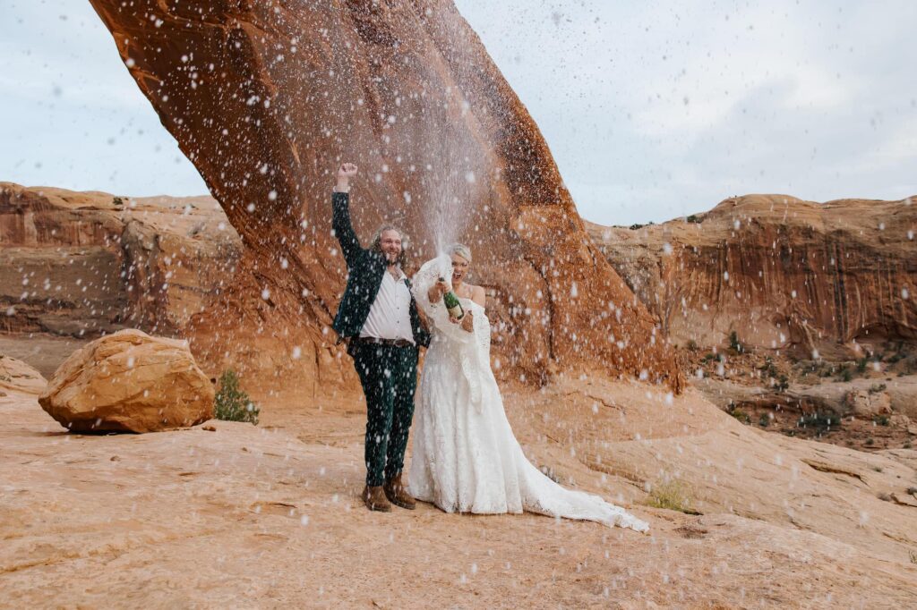 A bride and groom cheer and celebrate their elopement by popping a bottle of champagne. They are standing in front of the Corona Arch in Moab