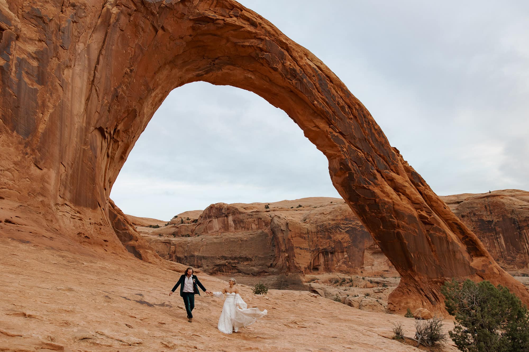 A bride and groom walk hand in hand on their elopement day in front of the Corona Arch in Moab