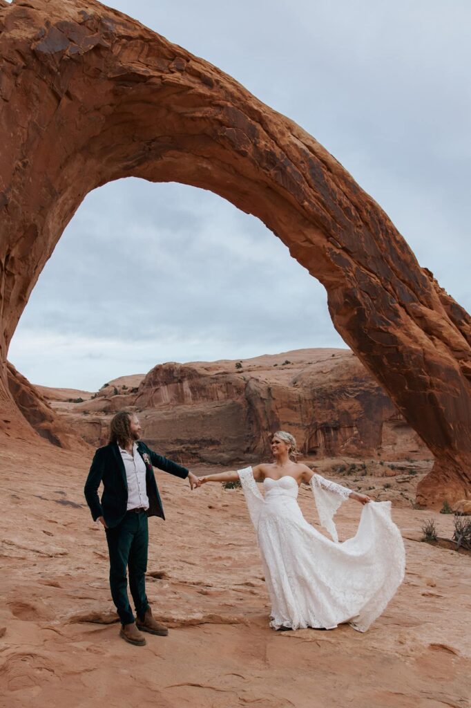 A bride and groom hold hands in front of the Corona Arch. The bride is holding her dress train