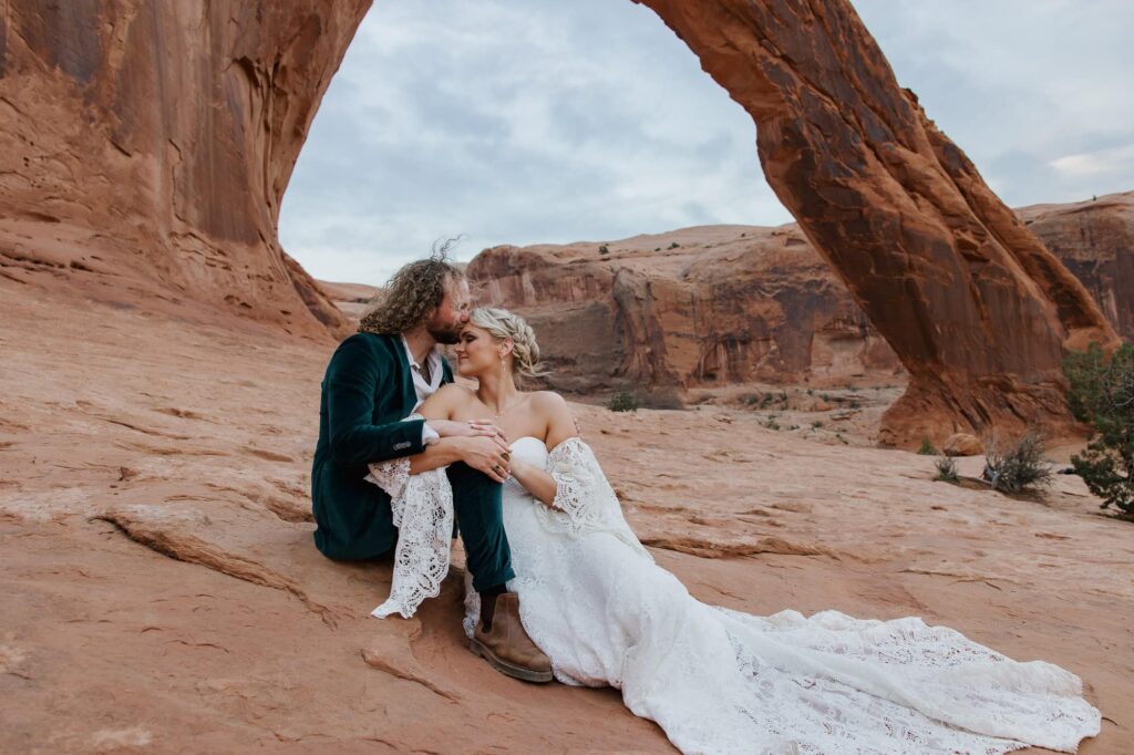 A bride and groom sit under the Corona Arch in Moan Utah during their sunset adventure elopement. 