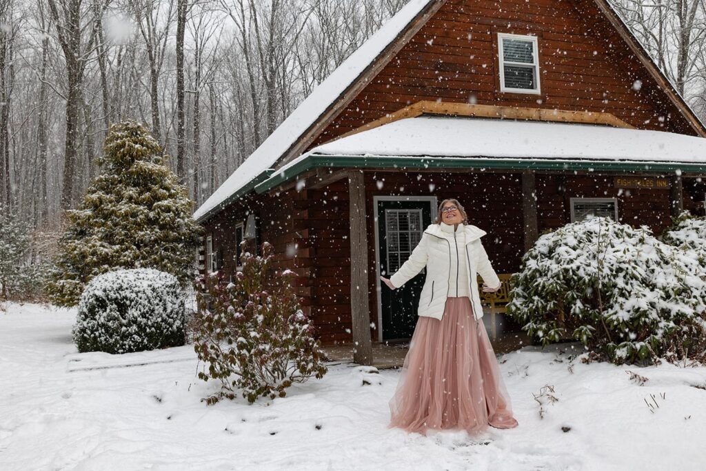 A bride, visually ecstatic by snow on her wedding day stands looking up at the sky in front of a cozy cabin near Ricketts Glen in Pennsylvania.