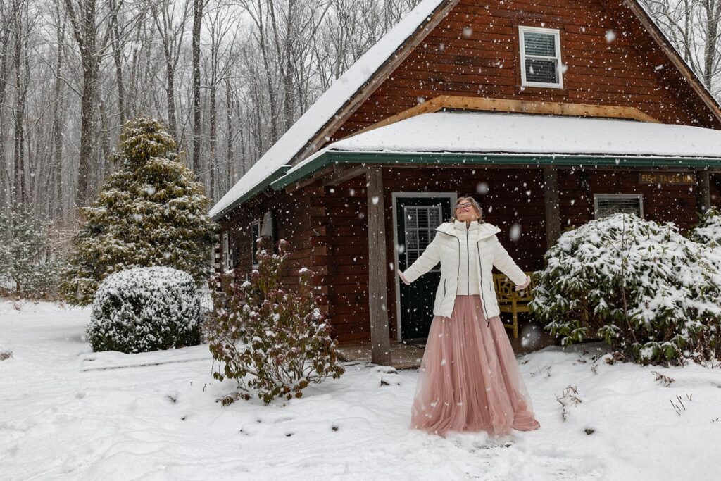 A bride, visually ecstatic by snow on her wedding day stands looking up at the sky in front of a cozy cabin near Ricketts Glen in Pennsylvania.