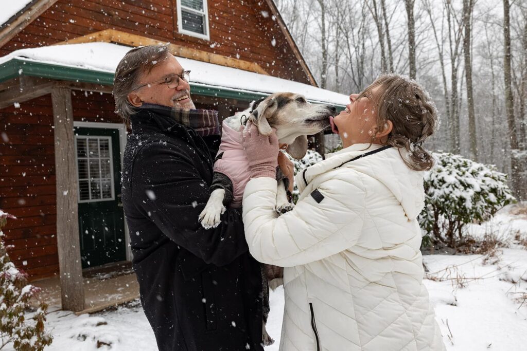 A bride and groom hold up their dog, as it kisses the brides face. They are standing in front of a cozy cabin, and it is snowing