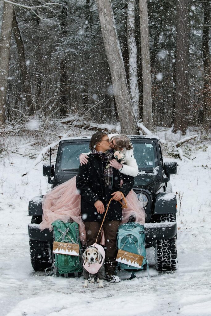 A bride sit on her jeep and leans on to her groom that is sitting in front of her. He also has their dog on a leash in front of them. It is snowing huge snowflakes, and it looks like they are in a winter wonderland.