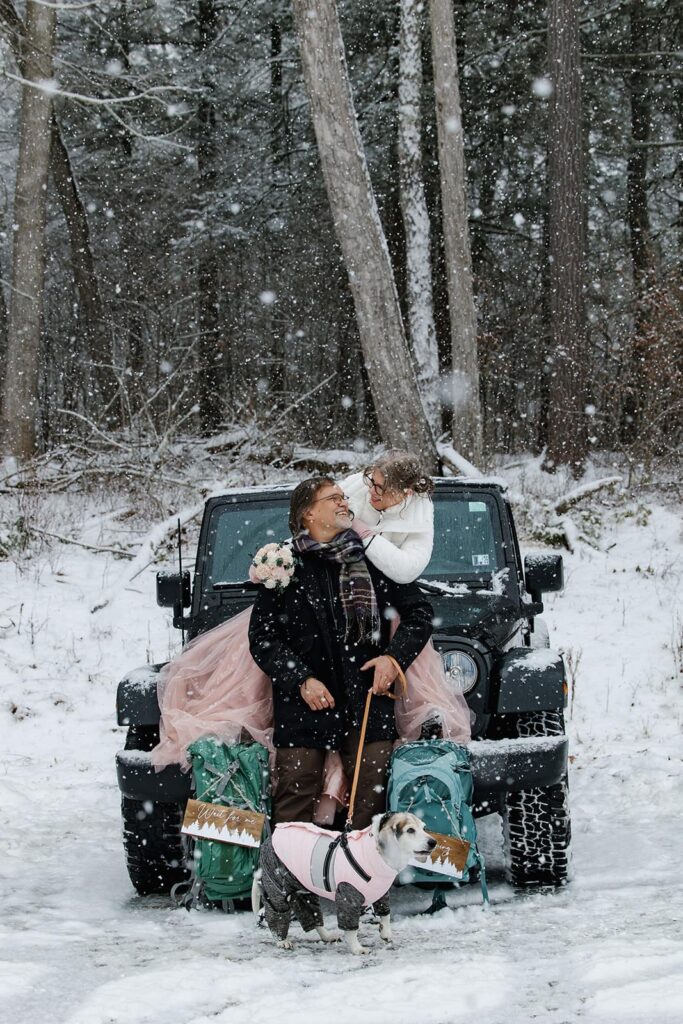 A bride sit on her jeep and leans on to her groom that is sitting in front of her. He also has their dog on a leash in front of them. It is snowing huge snowflakes, and it looks like they are in a winter wonderland.