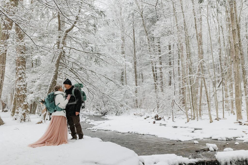 A bride and groom wearing hiking backpacks stand in what looks like a winter wonderland in Ricketts Glen. The bride is gently holding the cheeks of her now groom right before they kiss