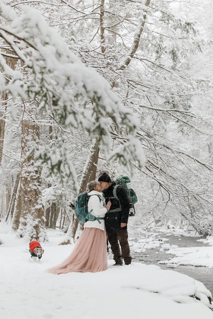 A bride and groom wearing hiking backpacks stand in what looks like a winter wonderland in Ricketts Glen by Kitchen Creek. The couple are sharing a kiss, and their dog is walking towrads them