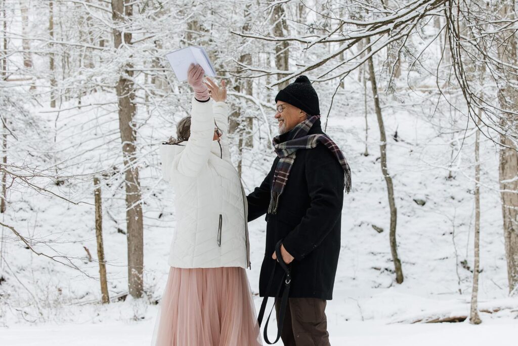 A bride lifts up her arms in celebration of her and her groom officially being married. They are surrounded by snow covered trees