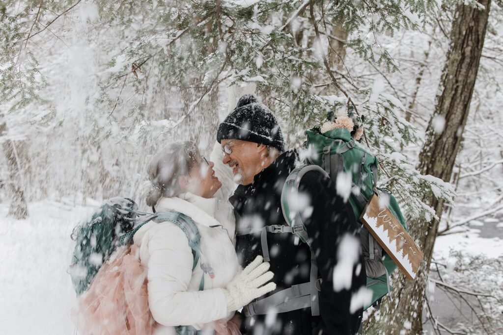 Snow falls on a bride and groom as they stand smiling at one another. 