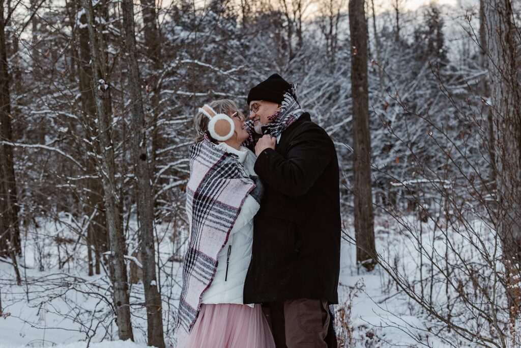 A bride and groom wrap up together in a cozy blanket and their winter clothing as they celebrate their wedding day in the snow.