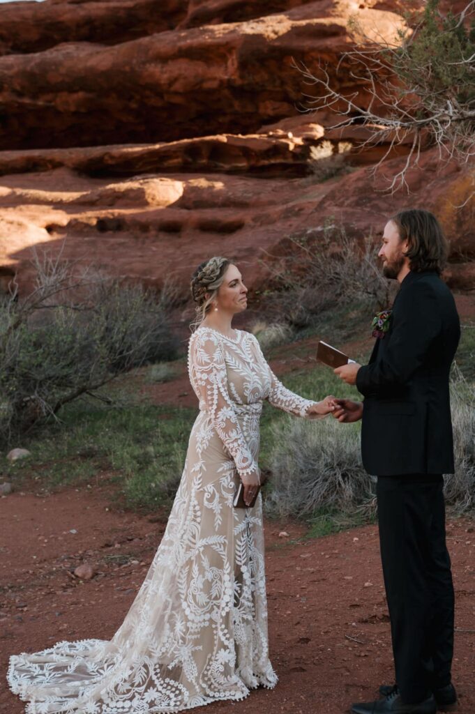 A groom reads his vows to his bride during their Moab elopement day. The bride is holding the grooms hand, and is wearing a see through lace dress with a boho flair