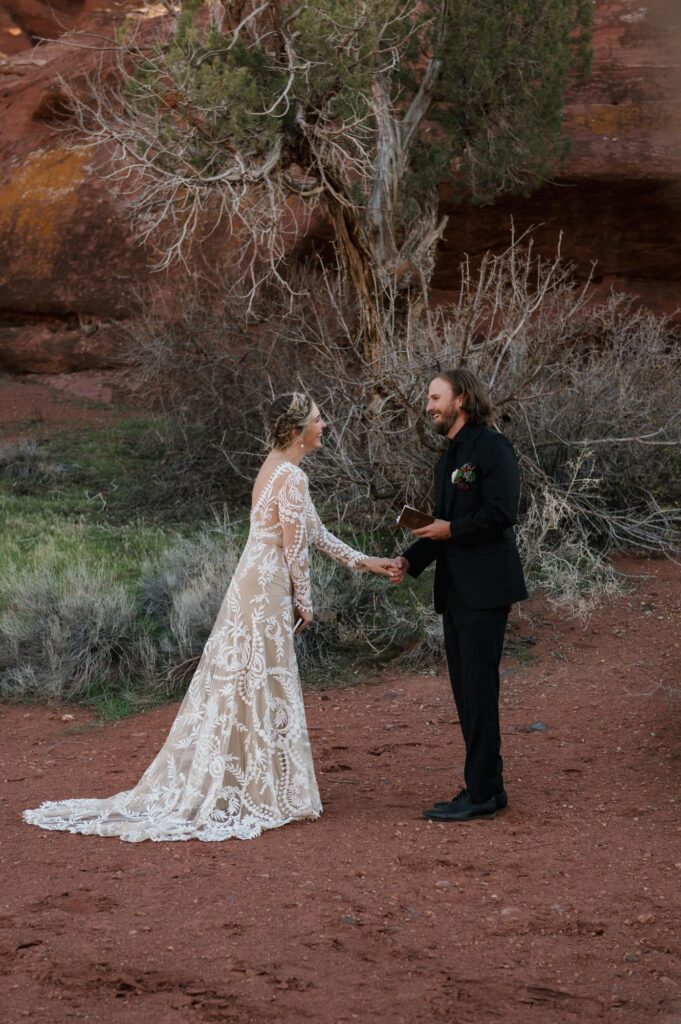 A bride and groom laugh during the vows of their Moab elopement day. The bride is holding the grooms hand, and is wearing a see through lace dress with a boho flair