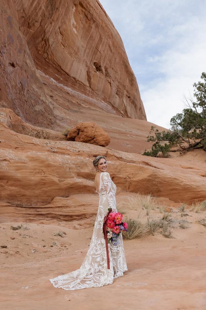A bride stands in front of the red rocks in her wedding dress holding her bouquet. She is looking over her shoulder and smiling.