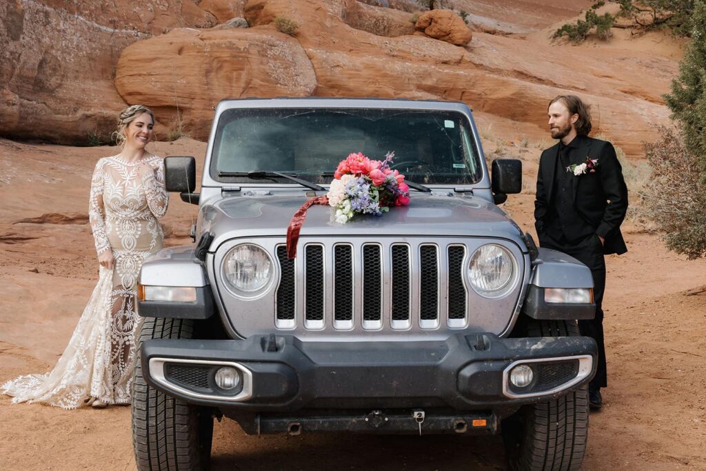 A bride and groom each putting on their final touches for their wedding day on either side of their jeep. The brides bouquet is sitting on the hood of the jeep. There are red rocks all around them. 