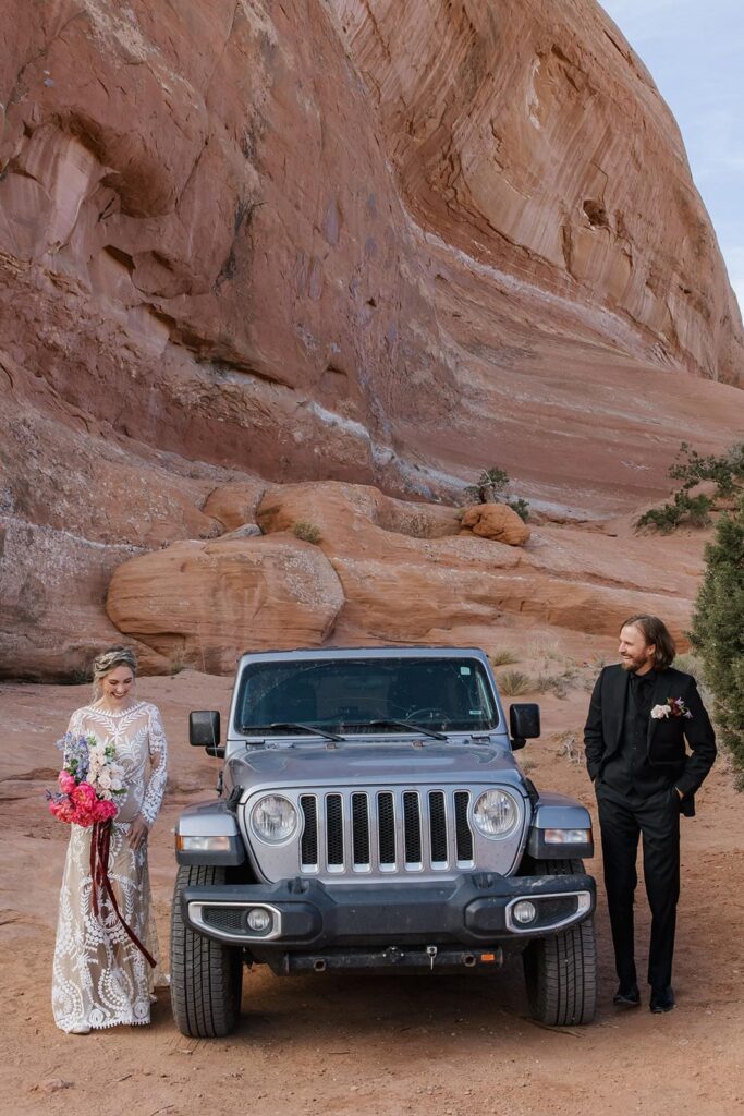 A bride and groom peek out from either side of a their jeep so they can see each other for the first time on their elopement day. They are standing in front of the red rocks and blur skies of Moab. The bride is wearing a see through lace dresws, and the groom is wearing an all black suit