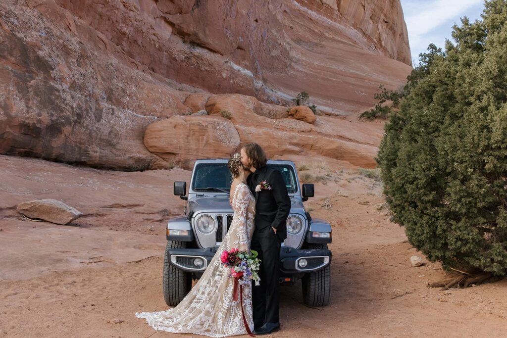 A bride and groom embrace in front of their jeep after the first look on their elopement day in Moab. they are standing in front of red rocks, and the bride is holding a colorful bouquet