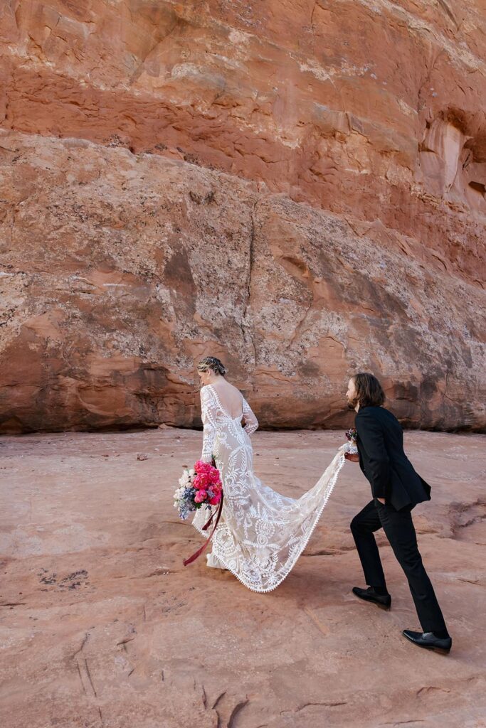 A groom carries the train of his brides dress as they walk up some red rocks on their way to Looking Glass Arch in Moab