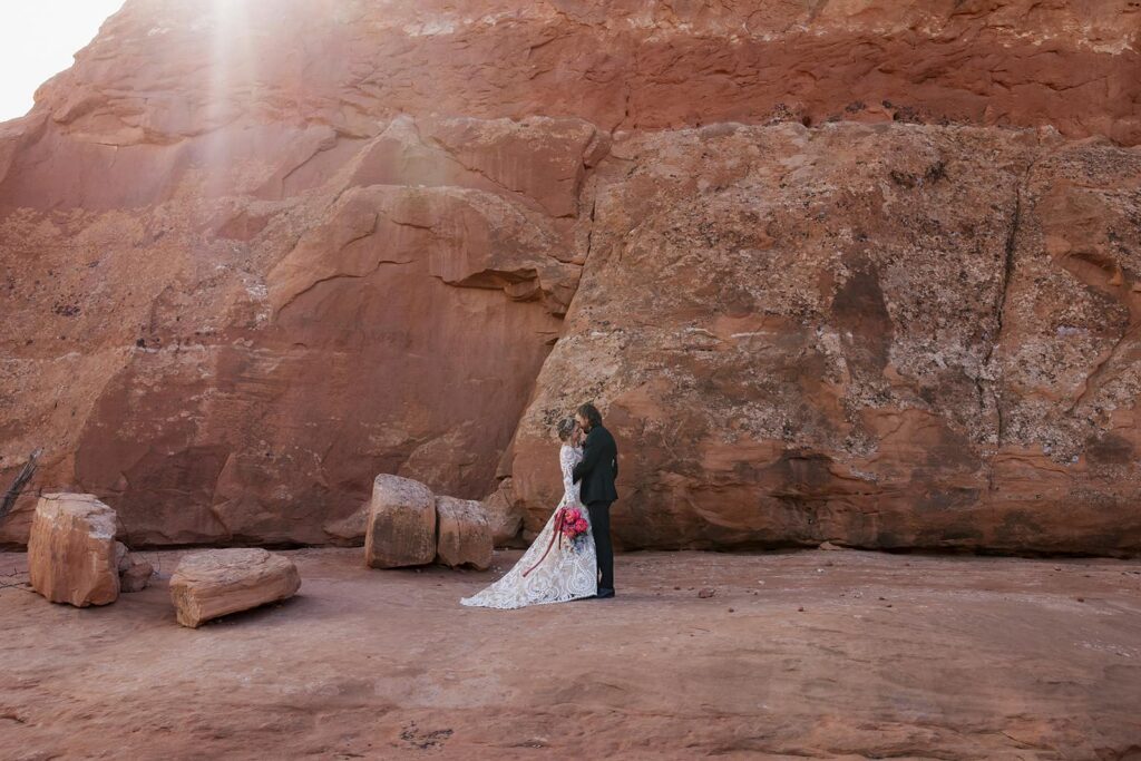 A groom kisses his brides forehead as they stand in front of red rocks as the sun flares behind them, in Moab. The bride is smiling, and holding her bouquet 