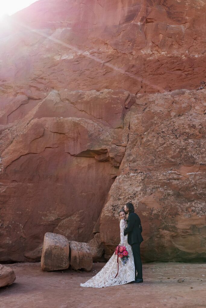 A groom kisses his brides cheek as they stand in front of red rocks as the sun flares behind them, in Moab. The bride is smiling, and holding her bouquet 