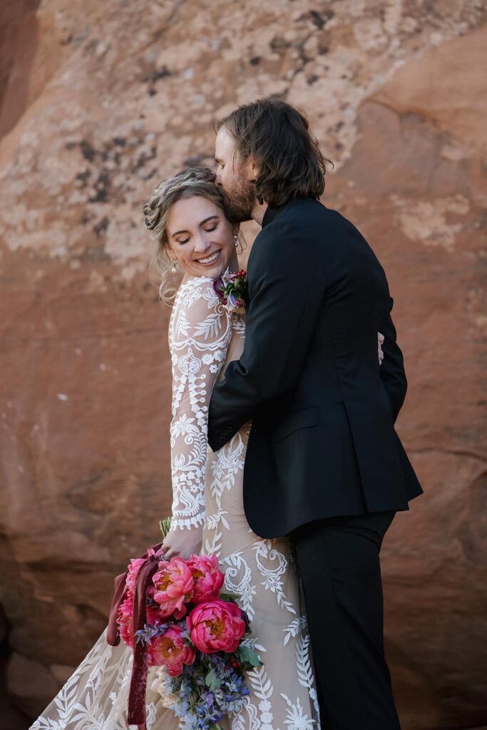 A groom kisses his brides cheek as they stand in front of red rocks in Moab. The bride is smiling, and holding her bouquet 