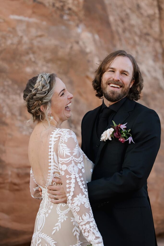 A bride and groom embrace, but both are laughing. The bride is wearing a backless see through lace dress and the groom is wearing all black. They are standing in front of the red rocks of Utah
