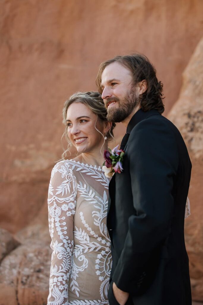 A bride and groom look off to the side smiling. They are standing in front of red rocks in Moab on their elopement day.