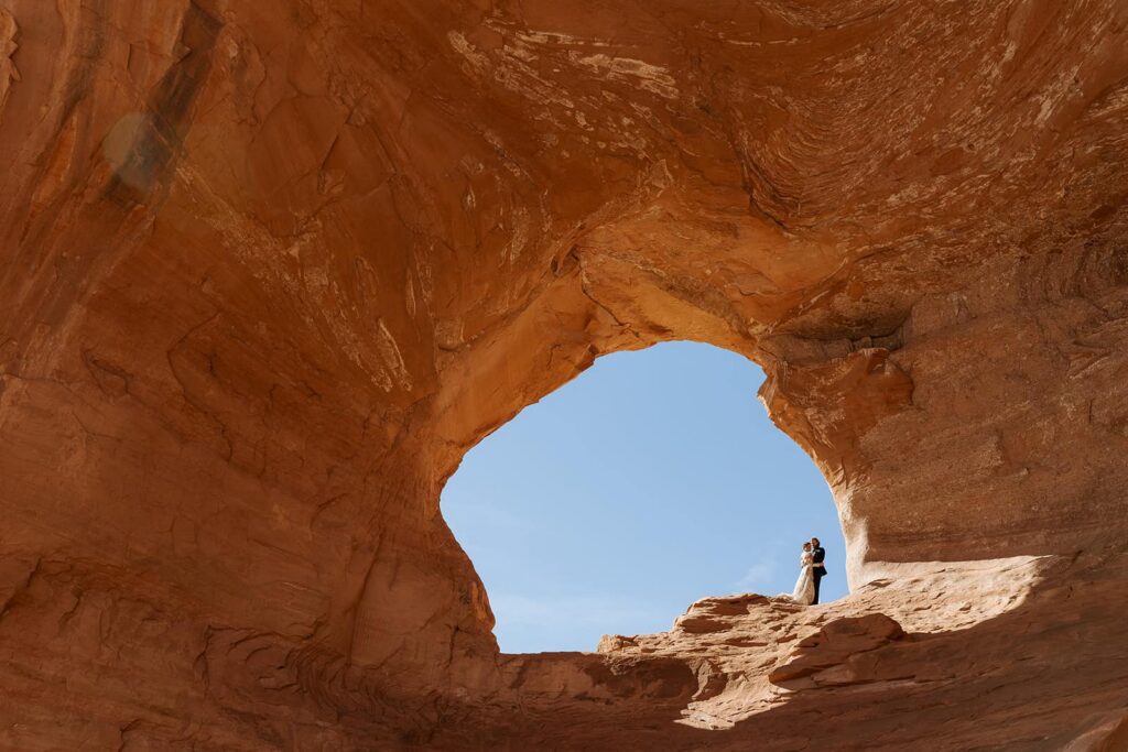 A bride and groom stand in the middle of Looking Glass Arch in Moab during their adventure filled elopement. The couple is embracing with blue skies behind them