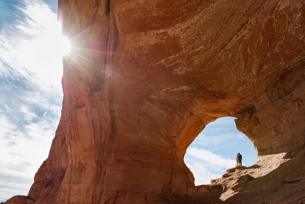 A bride and groom stand in the center of Looking Glass Arch with the blue sky behind them during their elopement in Moab. The sun is also flaring in the corner