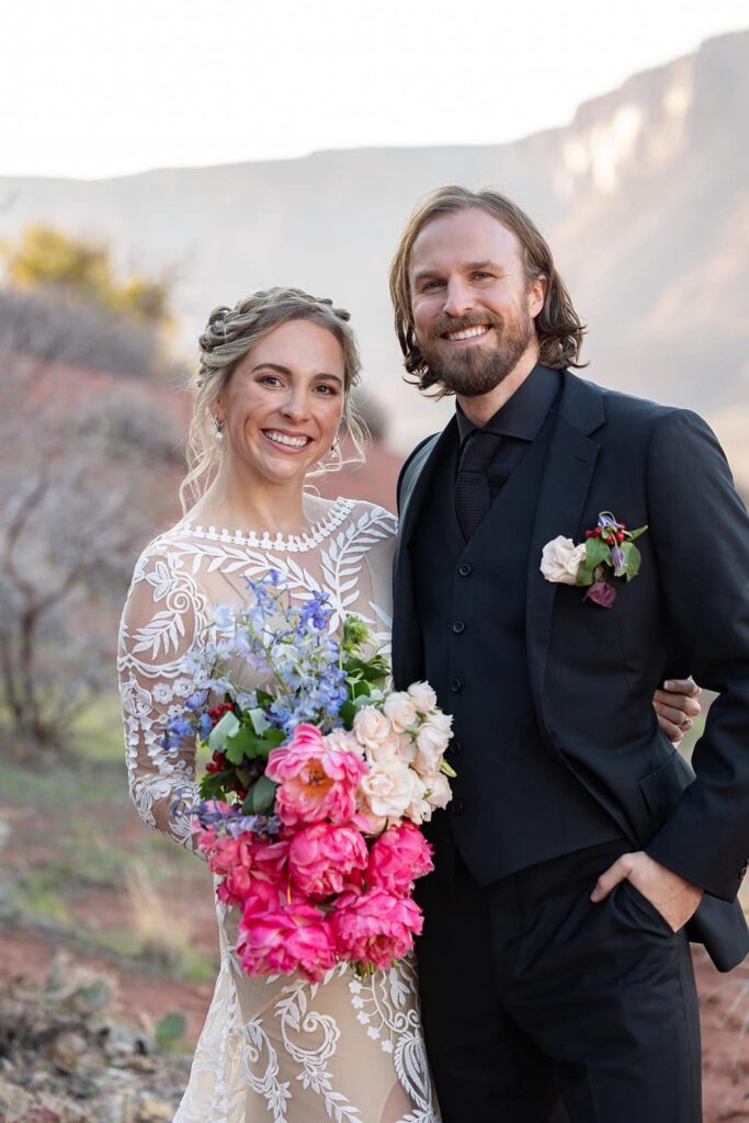 A bride and groom slie at the camera on their wedding day. The bride is wearing a sheer lace wedding dress and the groom is wearing all black. The bride is holding a colorful bouquet