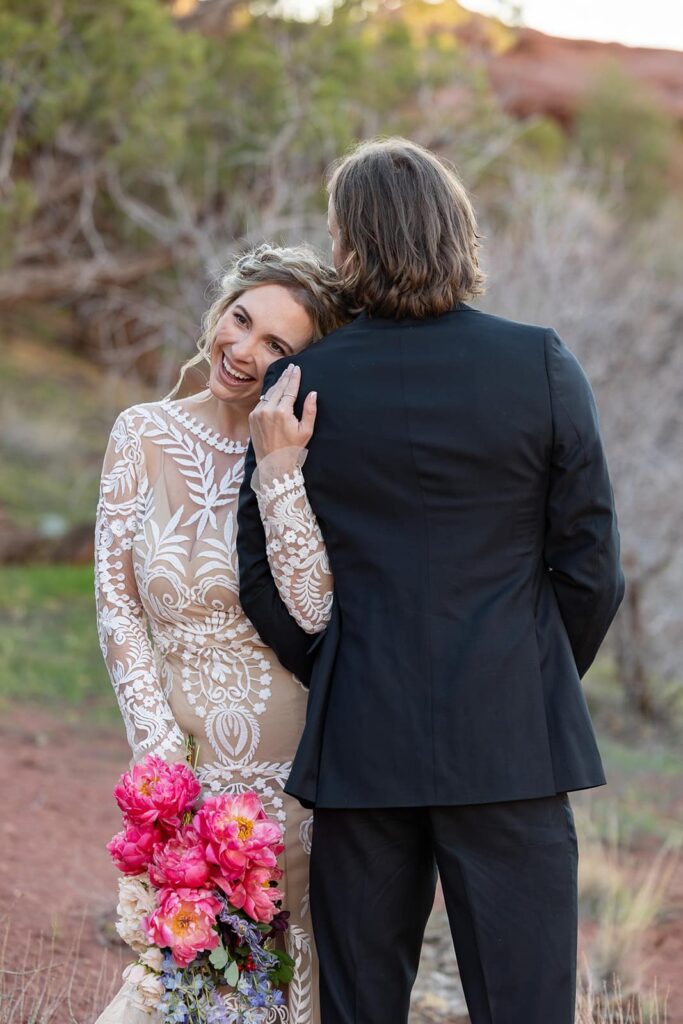 A bride faces the camera as the groom is turned around the other way. The bride is holding a pink bouquet and smiling,