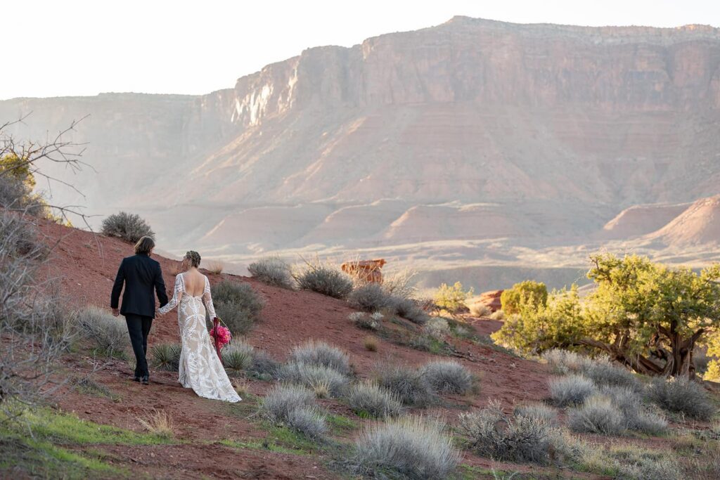 A bride and groom walk while holding hands through Castle Valley as the sunsets in Castle Valley. The bride is wearing a sheer lace dress and the groom is wearing an all balck suit. 