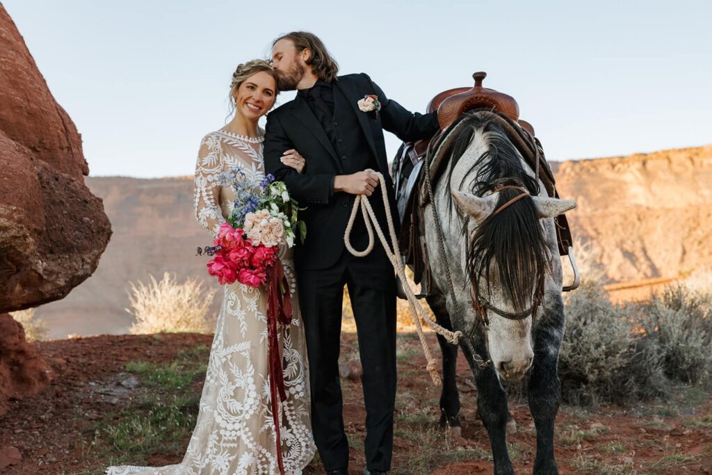 A groom kisses the head of his bride while she smiles. He is holding the reigns of a horse, and they are standing in Castle Valley in Moab