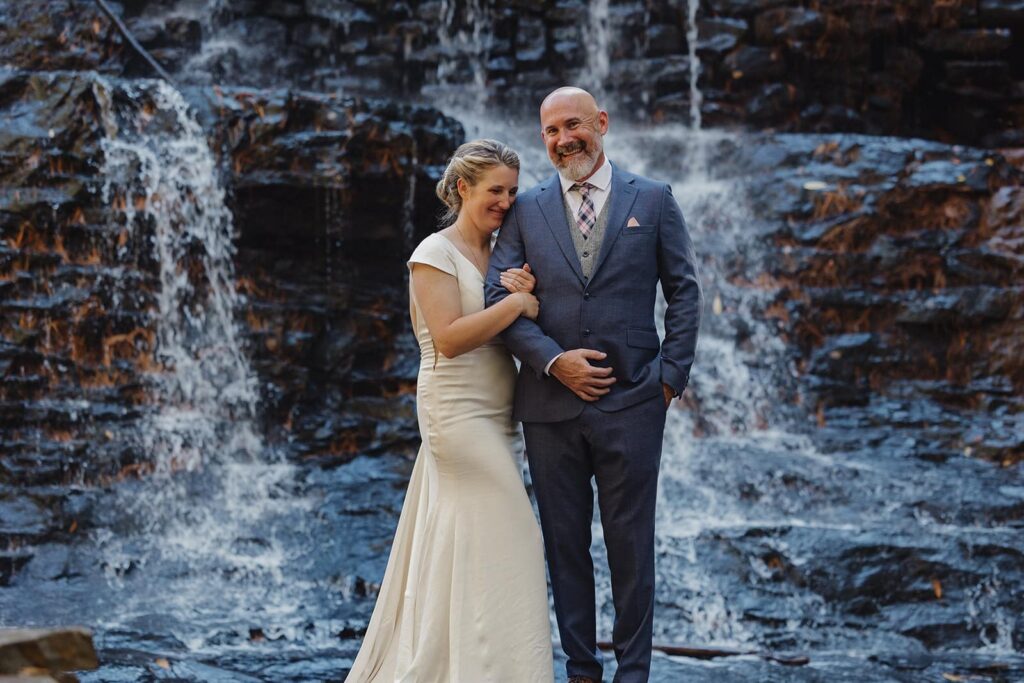 A bride leans into her husbands shoulder laughing as they stand in front of a waterfal at Hickory Run State Park during their intimate wedding