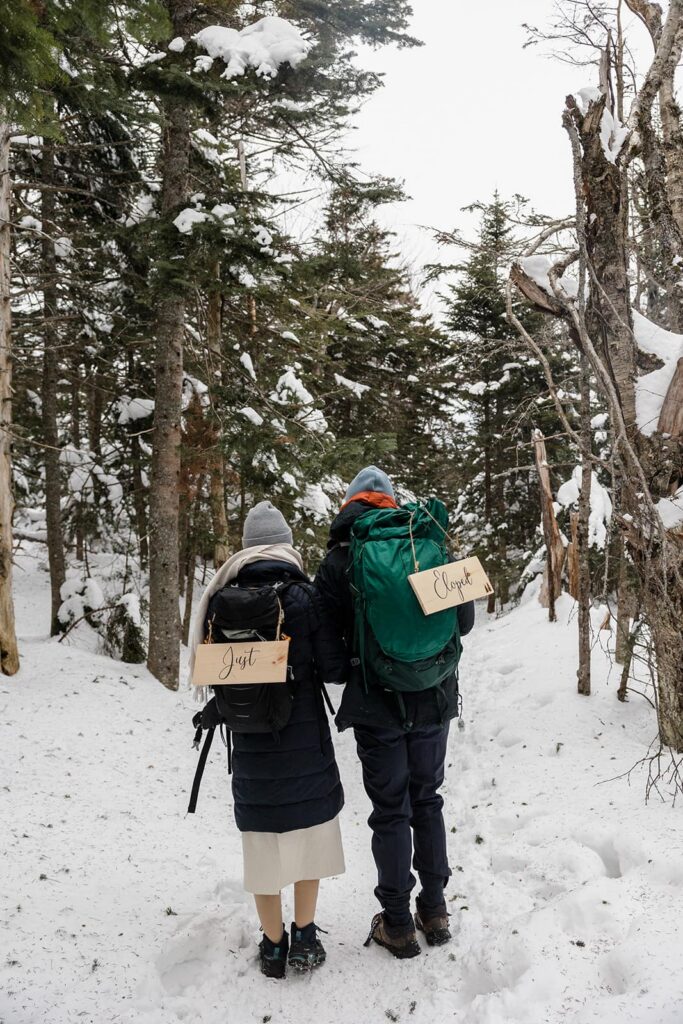 A bride and groom hike in the snow on their elopement day. They are both wearing backpacks  and have signs that say just eloped