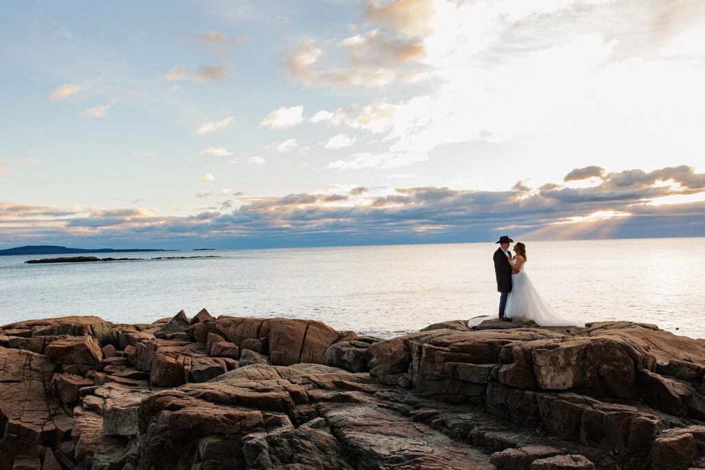 A bride and groom stand on the. liffs at Acadia National Park facing one another as the sun rises
