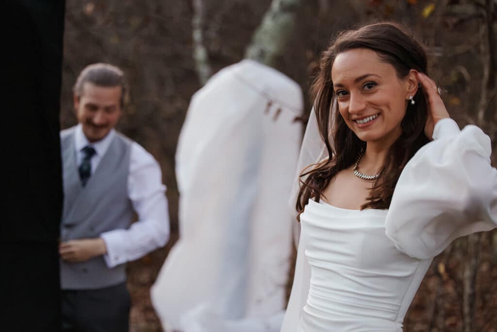 A bride smiling at the camera and tucking her hair behind her ear while her groom finishes getting dressed in the background at stony man